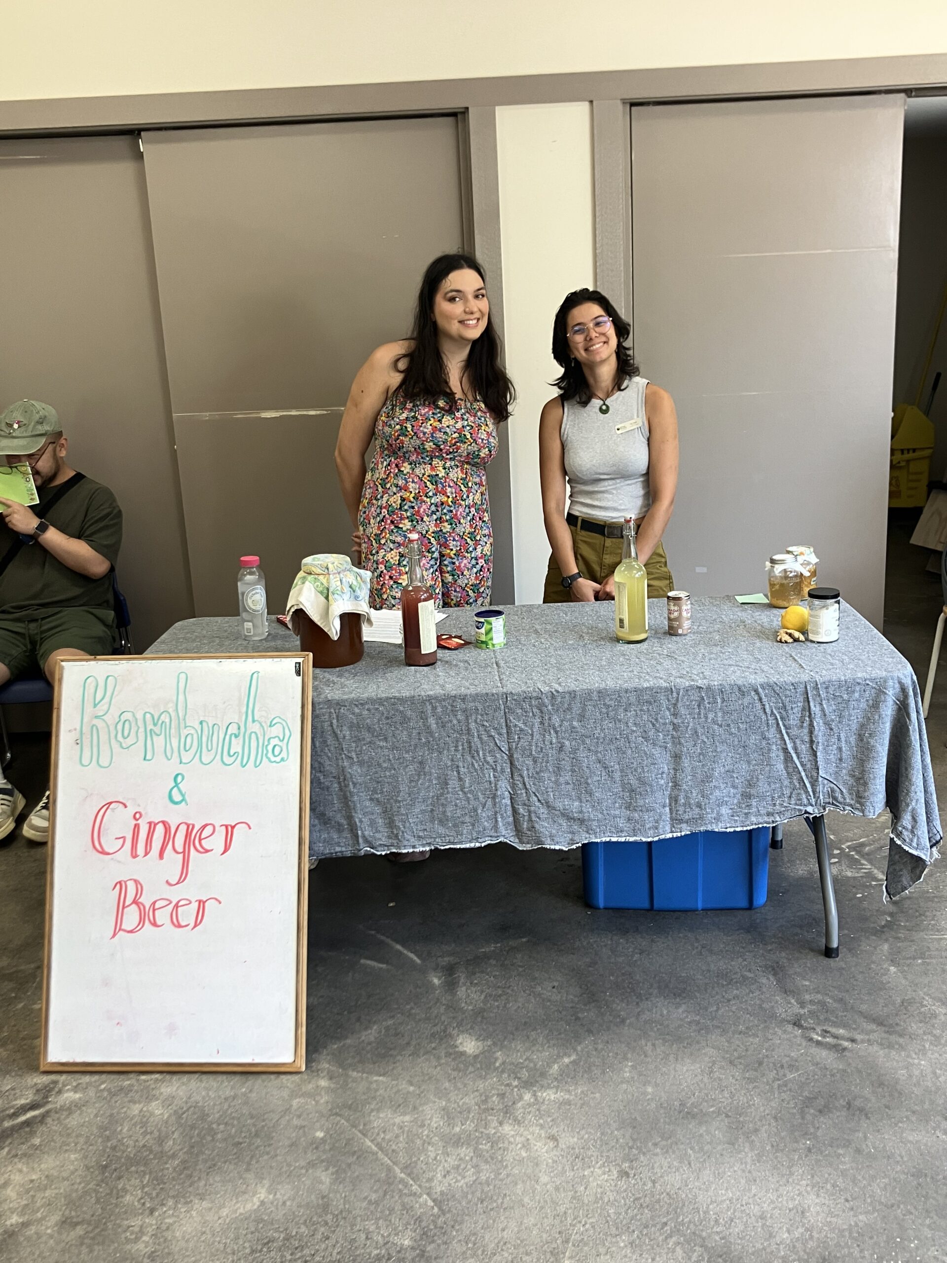 Kat (Urban Bounty Events and Volunteer Coordinator) and Gloria (UBC Nutrition Master's Student) pose in front of a table with kombucha and ginger beer. 
